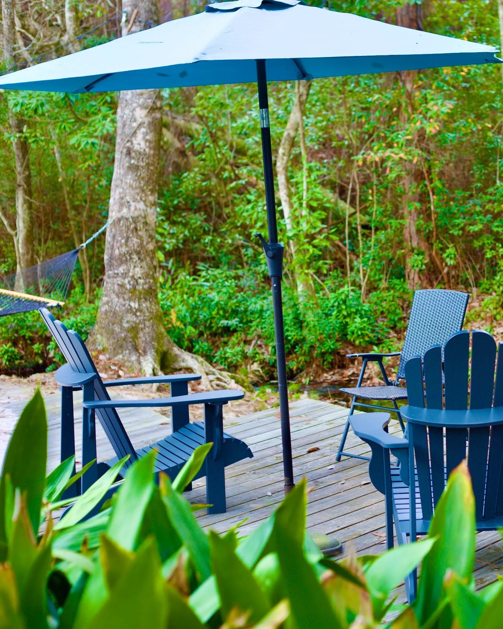 Outdoor seating area with blue Adirondack chairs and an umbrella beside the woods